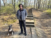 Performing and recording artist Bill Nace standing at the base of a trail surrounded by trees and next to a dog