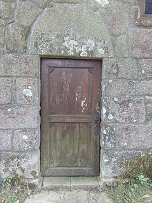 A wooden door on a granite wall. The top of the door is inscribed "ES YGLESIA DE REFUGIO".