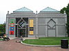 A one-story symmetrical granite building with two green peaked roofs either side of center. The entrance door is on the left and a similar design is on the right made out of a slightly darker granite.