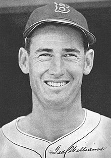 A man wearing a baseball hat, which has a "B" sewn onto it, smiles.