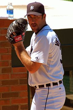 A light-skinned man with a goatee and wearing a white baseball uniform looks over his left shoulder while holding his right hand inside his black baseball glove.