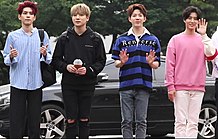 Four men standing outdoors in front of a black car, dressed casually in different styles.