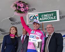 De trois-quarts, trois personnes sourient. Devant, un coureur au maillot rose lève un bouquet et tient un trophée.