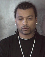 A photo of a man seen from the chest up. He has cornrows, a connected goatee and mustache, a tattoo on the left side of his neck, stud earrings, a chain necklace, and is wearing a black T-shirt.