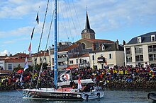 Le voilier descend le chenal des Sables-d'Olonne. Foule considérable sur le quai. Église en arrière-plan.