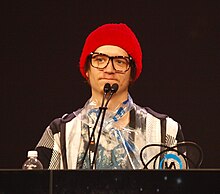 A 25-year-old Swedish man in a light grey jacket and a red bonnet speaking at a conference.