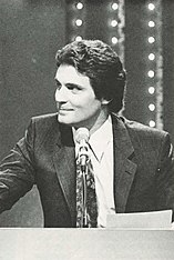 Host Robin Ward, a white man with dark hair wearing a suit, in front of a microphone and podium with marquee lights behind him