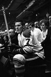 Tim Horton sitting on the bench during an ice hockey game with several other teammates.