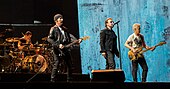 Four men on a stage, all wearing black clothing. Two are playing guitars, one is sitting behind a drum set, and one is singing into a microphone stand.