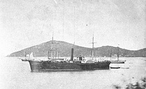 A black-and-white lithograph of the steamship USS De Soto, anchored in the harbor of Ponce, Puerto Rico. De Soto is in the foreground, facing left. De Soto is painted black or a very dark color, with the exception of her upper sidewheel guard which is white or very light in color. The ship has fine sweeping lines, a neatly rounded stern and slight clipper bow. She has two square-rigged masts with no sails, a tall, slim, raking smokestack amidships, just forward of the paddlewheels, and no apparent superstructure apart from what looks like temporarily rigged awnings on the main deck. The ship's walking beam engine can clearly be seen rising between the paddlewheels. In the distance are three sailing ships, one with a military appearance, and behind them two rounded, tree-covered hills which look almost uninhabited.
