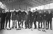 Black and white photo of eight hockey players wearing equipment and uniforms standing on an ice rink, along with four men wearing suits and full-length overcoats