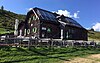 Schrägaufnahme einer aus Stein erbauten Berghütte mit Solarzellen auf dem Dach; um die Hütte ein kleiner Holzzaun; hinter der Hütte ein grasbewachsener Hang