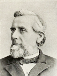 Head and shoulders photograph of a man in an overcoat. He has a salt and pepper goatee and has his hair combed to the side. He wears a calm expression and is looking slightly to the right.