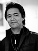 A black and white photo of a smiling Japanese man with short black hair and a black, zipped jacket shown from the shoulders up. The background is fuzzy and difficult to distinguish.