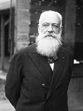 Portrait en buste d'un homme portant un costume complet et une barbe blanche, les bras croisés dans le dos.