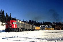 A passenger train exiting a mountain snowshed. The four diesel locomotives are dark gray, with a red nose on the lead locomotive. Four passenger cars are visible - one dark grey and three yellow.