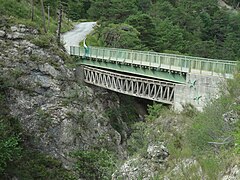 Un pont en croisillons de bois franchit un ravin, remplacé par un autre pont à poutre métallique construit juste au-dessus.