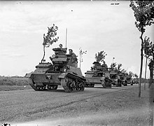 Photograph of several tanks and a truck driving along a road