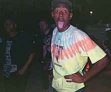 A flash in the dark lights up a man with a striped tie-dye t-shirt, arms akimbo, making a funny face with eyes wide and mouth open.