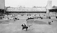 Photo en noir et blanc représentant l'enceinte du stade avec ses gradins; des obstacles étant montés sur la piste et un cavalier au galop au premier plan s'apprêtant à les franchir.