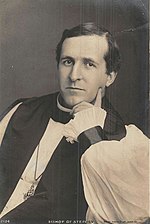 Black-and-white photo of a dark-haired white man, seated and resting his left hand around his chin, dressed in bishop's robes (rochet and chimere, pectoral cross and clerical shirt)