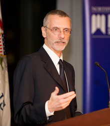 Side picture of Gius (man with glasses) giving a speech from a podium