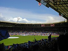 A sports stadium full of spectators. Those nearest the camera are waving blue and white flags.