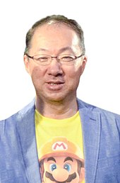 A Japanese man in his 50s. He has short brown hair and glasses and is smiling towards the camera. He is wearing a light blue blazer and a yellow shirt with Mario on it underneath.