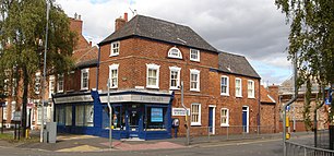 The corner of a terraced suburban street. The lower storey is a corner shop, now advertising as a chiropractic clinic. The building is two storeys high, with some parts three storeys high. It was formerly Alfred Roberts's shop.