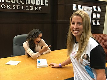 A young dark-haired woman sits at a desk in a bookstore, pointing to a blonde woman wearing her likeness on a t-shirt.