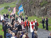 A crowd of people walking along an outdoor path. They are led by individuals in robes, and a number carry flag banners.