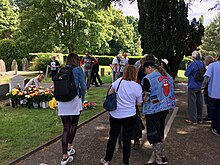People wearing clothes with Brian Jones and Rolling Stones fan art, stand by a grave stone decorated with many flowers in front. One person is placing a boquet.