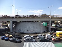 A road bridge spanning over an avenue with several vehicles. Below the bridge, there is a large cone-shaped air vent depicted.