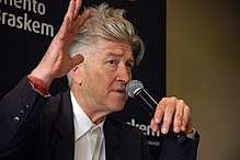 A man raises his hand and speaks into a microphone against a black backdrop.