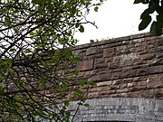 The bridge from the road, looking up through foliage at the east parapet wall with its unornamented date stone.