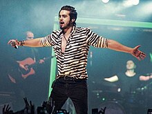 A dark haired man wearing a striped shirt extends his arms to the crowd while performing with a microphone