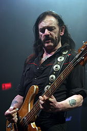 A man wearing a black shirt, looking down and playing a bass guitar.