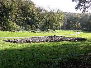 Rocks and boulders, forming a cairn, are retained by a short wall. Flat ground of short grass (the foreground in dappled shade and the rest in full sun) that surrounds the cairn is dissected by a pathway sweeping past and into the distance. In the background, trees, mainly in leaf, climb the steep sloped gorge.