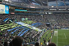 Fans waving flags and unfurling a large green and blue tifo behind a goal