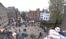 Wide shot of two barricades, as viewed from a rooftop behind the barricades. On this side of the barricades people can be seen painting "End Dutch complicity" on a banner. Protestors are gathered on the other side. Two police vans in the distance.
