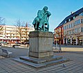 Fotoperspektive von vorne links auf das Denkmal, sodass der seitlich Sitzende Robert Schumann gut eingefangen wird. Die Skulptur ist grün angelaufen. Die Inschrift ist goldfarben. Auf den Stufen sind Aufkleber. Hinter dem Denkmal sind Sitzbänke, im Hintergrund kahle Bäume und parkende Autos.