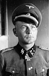 Black-and-white photo of a man wearing a peaked cap, military uniform with an Iron Cross displayed at his neck.