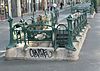 Photo en couleurs d'un entourage à cartouches ajouré vu du côté carré opposé à l'escalier et dont le socle en pierre s'orne de quelques tags