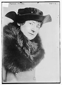 A white woman dressed in a dark fur wrap and a black hat with an upturned brim.