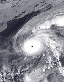 A photograph of an extremely powerful typhoon over the western Pacific Ocean. The circular eye is almost perfectly clear, and is surrounded by a large, nearly axisymmetric area of intense convection. There are two fragmented rainbands: one to the northeast, oriented horizontally; and the other to the east, oriented vertically. Arcs of thin, high clouds are present along the western, northwestern, and northern flanks of the typhoon.