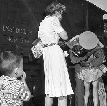 alt = A black-and-white photograph of a soldier's homecoming. The soldier is seen tightly hugging his 4–6 old daughter. His wife is stands shocked, covering her face with her hands. Their 2–3 year old son is standing in the corner, curiously observing the scene!