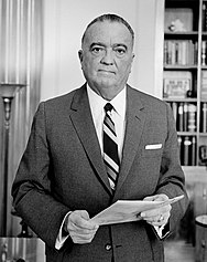 Hoover holding a file, looking at the camera; he wears a jacket and tie