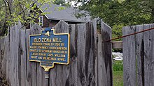 a picture of the marker, on a post like others but also attached to a fence through which you can see the stream that powered the mill