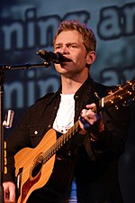 Chapman, holding an acoustic guitar, sings into a microphone on a stand.