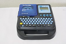 テプラの写真。本体の操作面は、記録部、液晶ディスプレイ、コンパクトにまとめられたキーボードの、3つで構成されている。本体と一体型の把手が付いている。